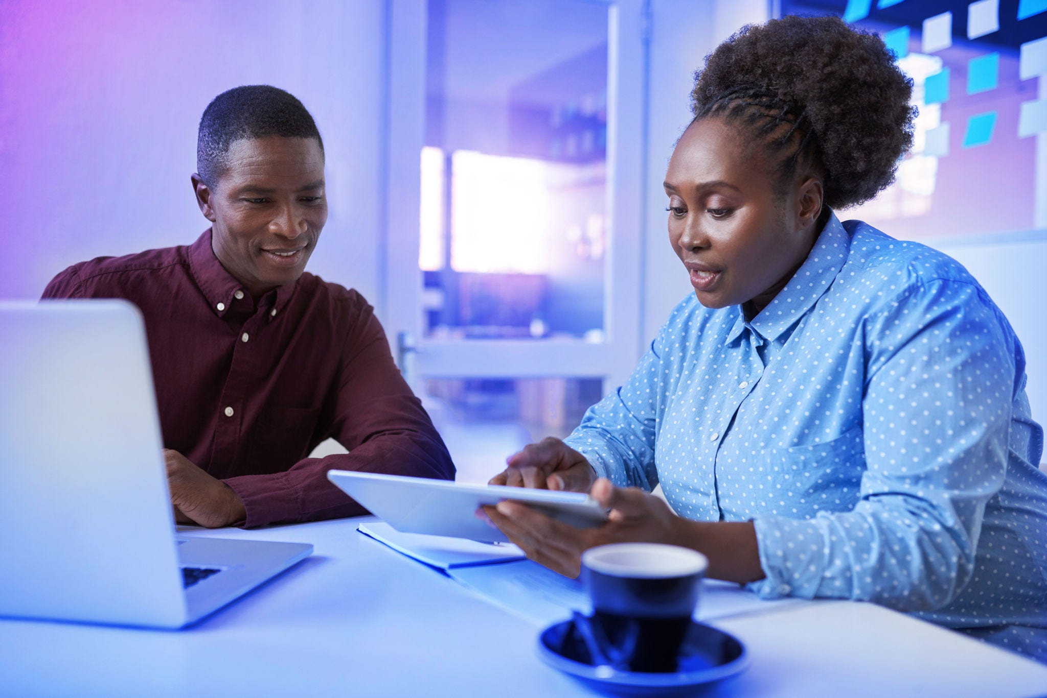Two casually dressed African colleagues sitting together at a table in a modern office working with a digital tablet and laptop
