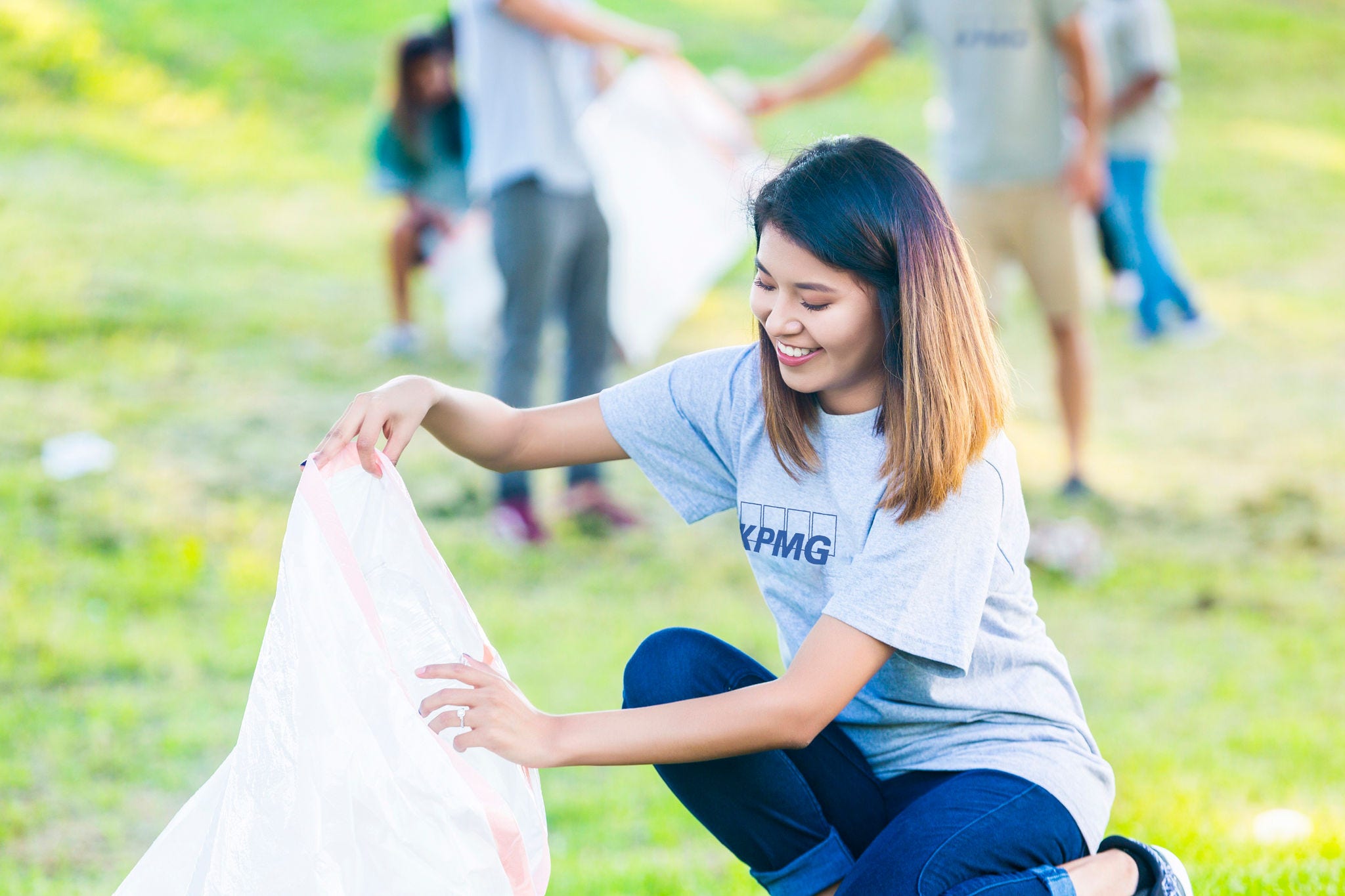Beaufiful young woman holds white trash bag while she helps her neighbors clean up thier park or neighborhood. She is kneeling down and wearing a gray volunteer t-shirt  and blue jeans. People are picking trash up in the background.